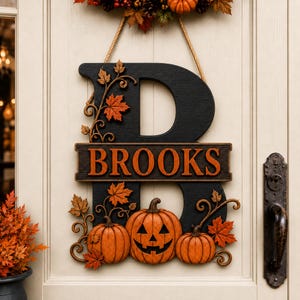May include: A black wooden letter B door sign featuring the name BROOKS in orange lettering. The sign is decorated with three orange pumpkins, including one jack-o'-lantern, and orange maple leaves with swirling vines, suspended by a rope.
