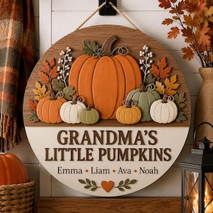 May include: A round wooden wall sign featuring a large orange pumpkin, smaller pumpkins in orange, white, and green, and autumn leaves. The text reads GRANDMA'S LITTLE PUMPKINS with the names Emma, Liam, Ava, and Noah printed below a small heart icon.