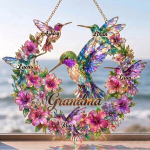 May include: A decorative hummingbird wreath with the word "Grandma" and the names "Ashley", "Ethan", "Lucas", "Liam", and "Mia". The wreath features colorful hummingbirds and flowers, suspended by a gold chain.