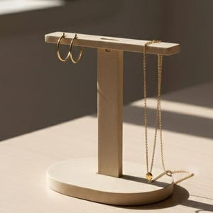 May include: A light beige jewelry display stand with a T-shape design. Gold hoop earrings hang on the top bar, and a gold necklace with a small pendant is displayed. The stand is on a light beige surface.