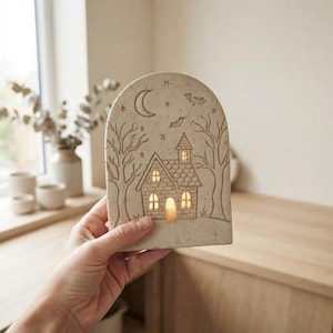 Op de afbeelding: Een beige keramische boogvormige lamp met een gegraveerde scène van een huis, kale bomen, een maansikkel, sterren en vleermuizen. De uitgesneden ramen en deur laten een warm licht schijnen dat afkomstig is van een interne lichtbron.