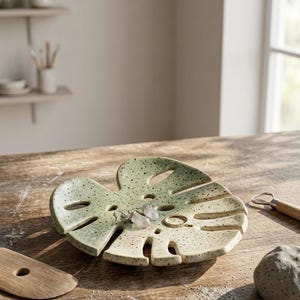May include: A ceramic trinket dish shaped like a monstera leaf with a speckled light green and cream glaze. The dish features cut-out leaf veins and holds a pair of clear crystal drop earrings and a thin gold-toned metal ring on its surface.