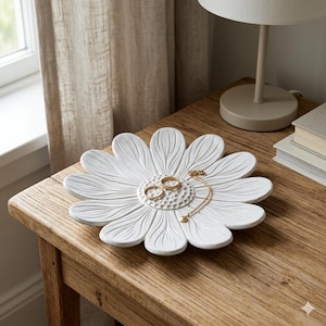 Peut inclure: Un vide-poche en céramique blanche en forme de fleur de marguerite avec des pétales texturés et un motif central en nid d'abeille. Deux bagues dorées et un collier à chaîne dorée délicat reposent au centre du plat à la finition mate.