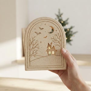 May include: An arched, speckled beige ceramic luminary featuring a cutout design of a house, bare trees, bats, and a crescent moon. The interior is illuminated, casting a warm glow through the windows of the house and the surrounding scene.