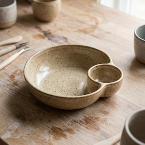 May include: A speckled beige ceramic bowl featuring a smaller integrated circular compartment on one side. The bowl has a smooth, glossy finish with visible dark brown flecks throughout the clay body, creating a textured appearance for this dish.