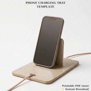 Può includere: Un vassoio di ricarica per telefono beige con un telefono in una custodia. Il vassoio ha una base rettangolare con un retro rialzato per tenere il telefono in posizione verticale. Il testo "PHONE CHARGING TRAY TEMPLATE" è in alto.