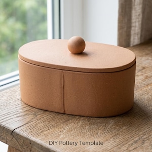 Könnte beinhalten: Eine terracottafarbene, ovale Keramikbox mit Deckel und kleinem kugelförmigem Griff. Die Box steht auf einer Holzoberfläche in der Nähe eines Fensters. Der Text "DIY Pottery Template" befindet sich am unteren Bildrand.