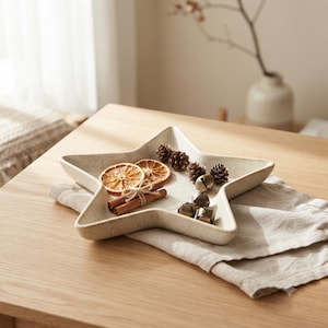 Peut inclure: Un plateau en céramique en forme d'étoile avec une finition crème mouchetée. Le plateau contient deux tranches d'orange séchées, un fagot de trois bâtons de cannelle liés avec de la ficelle, quatre petites pommes de pin et trois grelots dorés.