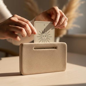 Puede incluir: Un tarjetero rectangular beige con una ranura en la parte superior, que contiene una carta de tarot. La carta presenta un diseño de sol con una luna creciente. El tarjetero está sobre una superficie clara.
