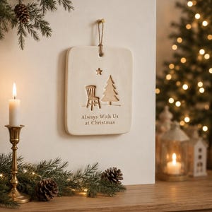 May include: A rectangular off-white ceramic wall plaque hanging from a jute string. The plaque features an embossed design of a chair, a Christmas tree, and a star, with the text Always With Us at Christmas printed in dark lettering.