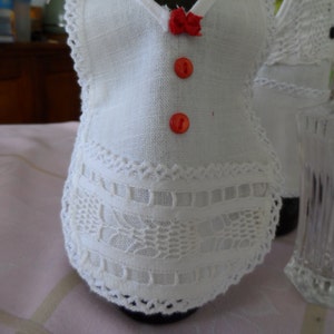 May include: White bottle cover with lace trim and red buttons. The cover is designed to look like a dress.