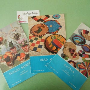 May include: Three vintage craft books with colourful covers. The first book is titled "Dualso Baskets" and features a photo of three woven baskets with flowers. The second book is titled "Bead Making" and features a photo of colourful beaded crafts. The third book is titled "Raffia Handicrafts" and features a photo of woven raffia crafts.