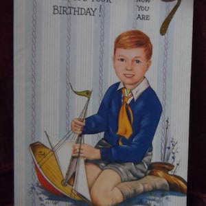 Op de afbeelding: Vintage verjaardagskaart met een jongen met rood haar, gekleed in een blauwe trui en korte broek, die een speelgoed zeilboot vasthoudt. De kaart heeft de tekst "Hurrah! It's Your Birthday!" en "Now You Are 7" op een gestreepte achtergrond.