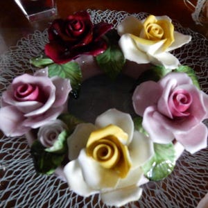 May include: A porcelain floral arrangement featuring five roses in shades of pink, yellow, and red. The roses are arranged in a circle and have green leaves. The arrangement is on a white lace background.