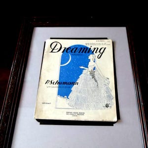 Könnte beinhalten: Ein Vintage-Notenblatt für das Lied "Dreaming" von R. Schumann. Das Cover zeigt eine Frau in einem weißen Kleid, die vor einem blauen Himmel mit Sternen und einer Mondsichel steht. Der Text "Dreaming" steht in großen, fetten Buchstaben oben auf dem Cover. Der Text "R. Schumann" steht in kleineren Buchstaben unter der Frau. Der Text "Banks Music House, 25 County Arcade, Leeds" steht unten auf dem Cover.