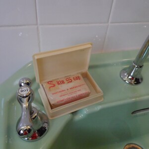 Może przedstawiać: Biała plastikowa mydelniczka z klapką na zawiasach, zawierająca mydło z etykietą "Skin Soap". Mydelniczka stoi na jasnozielonym porcelanowym zlewie z chromowanym kranem.