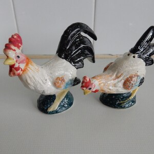 May include: A set of two ceramic rooster salt and pepper shakers. The rooster on the left is white with black, red, and blue accents. The rooster on the right is white with brown, black, and blue accents.