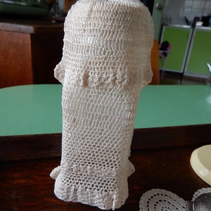 May include: Off-white, hand-crocheted bottle cover. The cover is cylindrical with a flared top and bottom, and features a delicate, openwork pattern. The crochet work is tightly woven, creating a textured surface. The cover is likely designed to fit over a bottle.