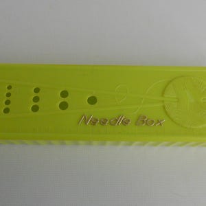 May include: A bright green plastic needle box with a gold inscription that reads "Needle Box". The box has a design of a ball of yarn and two needles.