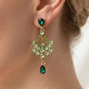 Puede incluir: Elegantes pendientes dorados con piedras preciosas verdes. Los pendientes presentan una gema en forma de lágrima en la parte inferior, un grupo de gemas más pequeñas en el centro y una gema más pequeña en la parte superior. Los pendientes están diseñados para colgar y captar la luz.