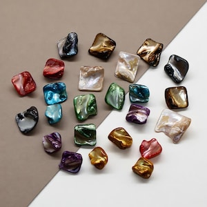 Op de afbeelding: Een verzameling iriserende, vierkante schelpkralen in diverse kleuren, waaronder rood, blauw, groen, goud en paars. De kralen hebben een natuurlijke, gepolijste uitstraling en liggen op een wit en bruin oppervlak. Elke kraal is ongeveer 1 cm.