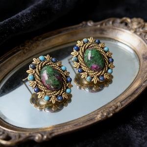 May include: A pair of stud earrings with oval-shaped green and red gemstones, surrounded by gold-toned metalwork and smaller blue and turquoise stones. The earrings are displayed on a vintage-style mirrored tray.
