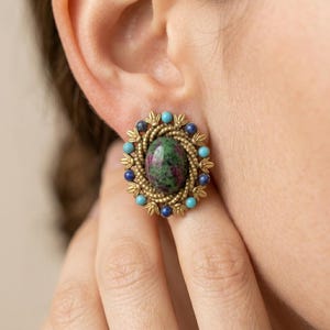 May include: A close-up of a gold-toned earring with a central oval stone featuring green and red hues. The earring is surrounded by small blue and turquoise beads, and a decorative gold-colored border.