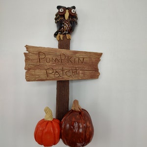 Pode incluir: Uma placa de madeira com as palavras "Pumpkin Patch" gravadas nela. Uma figura de coruja fica em cima da placa, e duas pequenas abóboras estão penduradas abaixo.