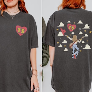 Graphic Comfort Colors t-shirt featuring a TS heart logo on the front and a concert-themed girl carrying a guitar with clouds and stars artwork on the back.