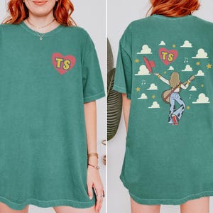 May include: A dark green short-sleeve t-shirt featuring a small red heart with yellow TS letters on the front. The back displays a graphic of a person with long hair playing a guitar, wearing cow-print pants, surrounded by clouds, stars, and a heart.