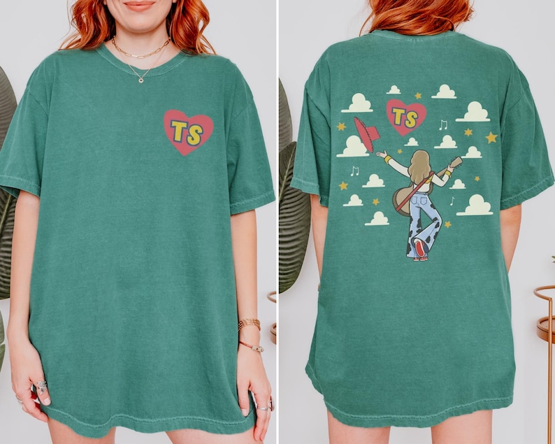 May include: A dark green short-sleeve t-shirt featuring a small red heart with yellow TS letters on the front. The back displays a graphic of a person with long hair playing a guitar, wearing cow-print pants, surrounded by clouds, stars, and a heart.