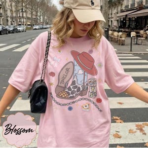 May include: A light pink t-shirt featuring a graphic print of a cowboy boot with a cow print pattern, a red hat, and blocks with the letters T, S, and the number 13. The design includes the name Taylor, a chain, flowers, and a bow.
