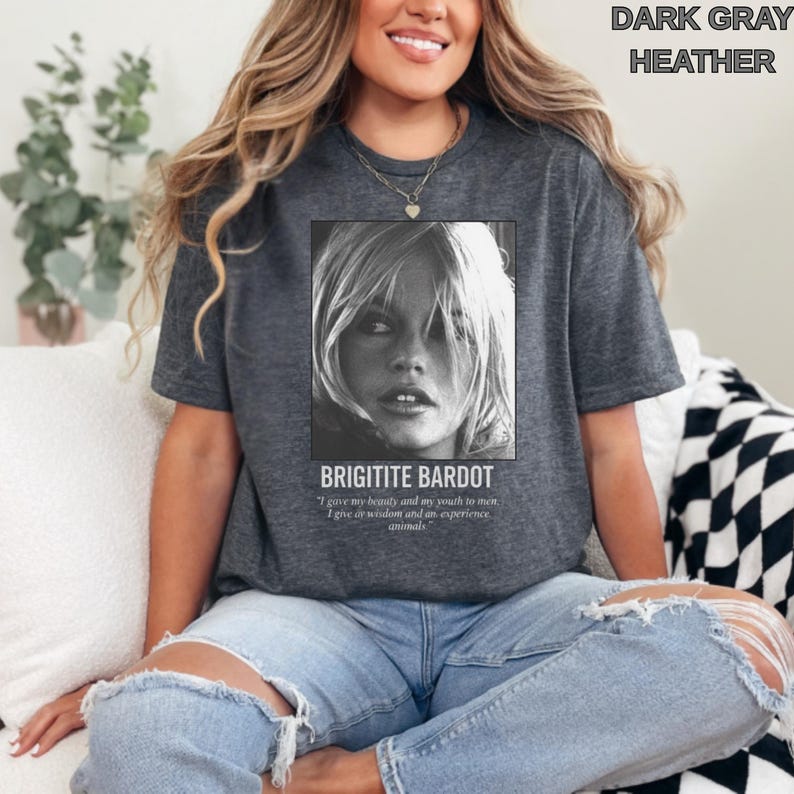 May include: Dark gray heather t-shirt featuring a black and white image of Brigitte Bardot. The text "BRIGITITE BARDOT" is below the image, along with a quote. The shirt is a casual style.