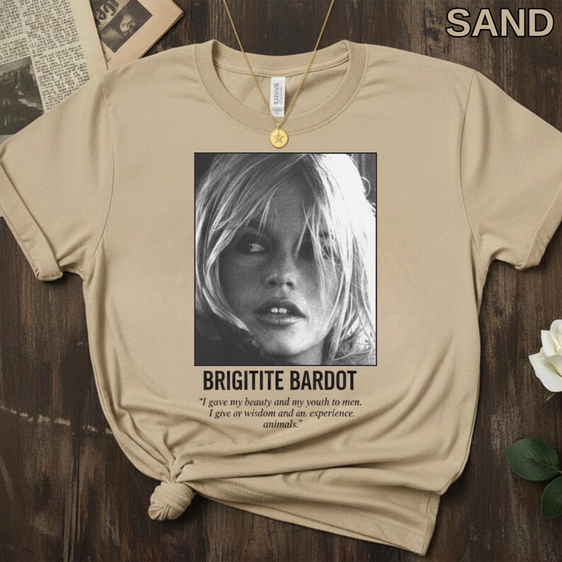 May include: Sand-colored t-shirt featuring a black and white image of Brigitte Bardot. The image is framed with the text "BRIGITITE BARDOT" and a quote about beauty, youth, wisdom, and animals. A gold necklace hangs on the shirt.
