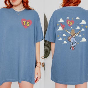 May include: A blue short-sleeve t-shirt featuring a red heart with the yellow letters TS on the front chest. The back displays a graphic of a person with long hair playing a guitar, a red hat, clouds, stars, musical notes, and a red heart with TS.
