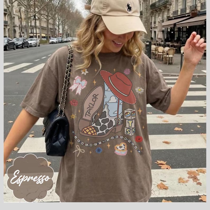 May include: A brown short-sleeve t-shirt featuring a graphic print of a cowboy boot with a cow print pattern, a red hat, a bow, and blocks with the letters T, S, and the number 13. The word Taylor is printed on the boot sole. A black quilted bag.