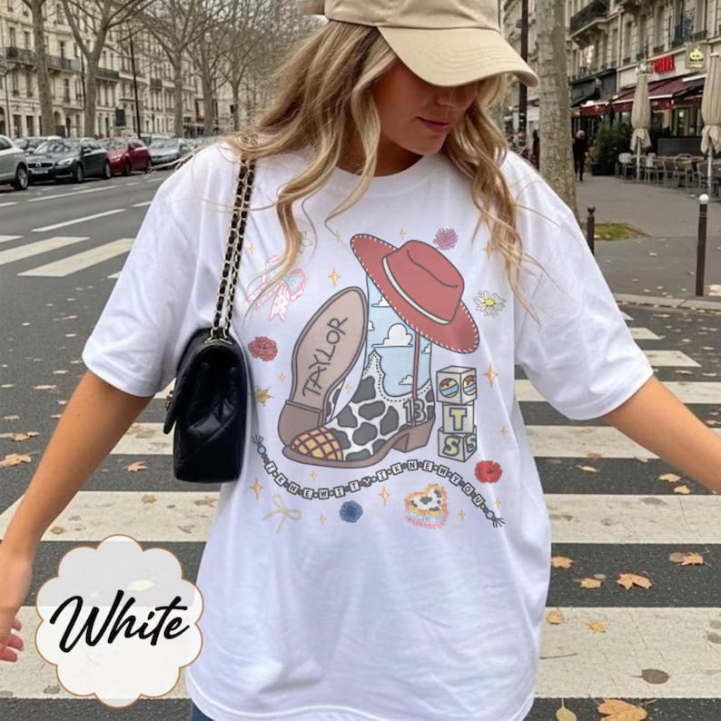 May include: A white t-shirt featuring a graphic print of a cow-print cowboy boot with the name Taylor, a red cowboy hat, stacked blocks with the letters T, S, and 13, small flowers, stars, and a beaded chain design with text along the bottom.