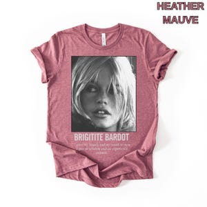May include: Heather mauve t-shirt featuring a black and white image of Brigitte Bardot. The image is framed with the text "BRIGITITE BARDOT" and a quote. The shirt is a solid color and has short sleeves.