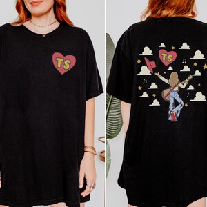 May include: A black t-shirt featuring a small red heart on the front left chest with the yellow letters TS inside. The back displays a graphic of a person with long hair holding a guitar and hat, surrounded by clouds, stars, and a heart with TS.