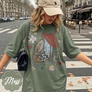 May include: An olive green t-shirt featuring a graphic print of a cowboy boot with a cow print pattern, a red hat, the name Taylor, the number 13, and stacked blocks with the letters T, S, and a heart. The design includes stars, bows, and flowers.