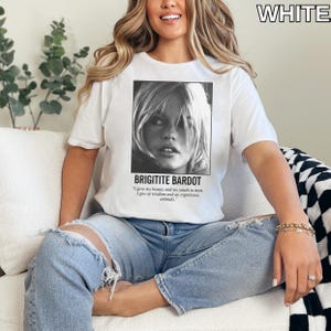 May include: White t-shirt featuring a black and white portrait of Brigitte Bardot. The text "BRIGITITE BARDOT" is below the image, along with a quote. The word "WHITE" is in the upper right corner. The shirt is worn with blue jeans.