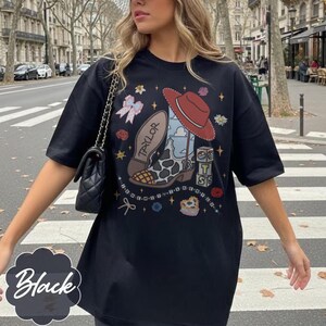 May include: A black short-sleeve t-shirt featuring a graphic print of a cowboy boot with a cow print pattern, a red hat, and blocks with the text TS. The boot has the name Taylor written on the sole. Small stars, flowers, and bows surround the central design.