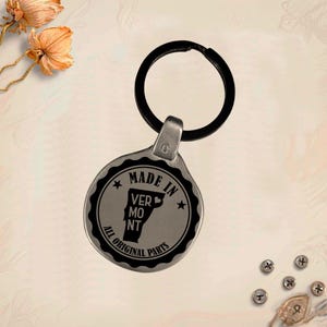 Può includere: Un portachiavi color argento con un portachiavi nero. Il portachiavi ha un design circolare con le parole "MADE IN VERMONT" e "ALL ORIGINAL PARTS". Lo stato del Vermont è al centro.