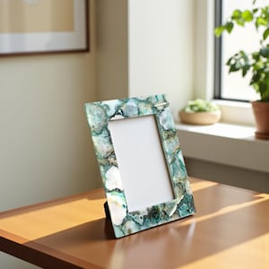 May include: A decorative picture frame with a rectangular opening. The frame is made of a mosaic of green, white, and gray stones. The frame is standing on a wooden surface.