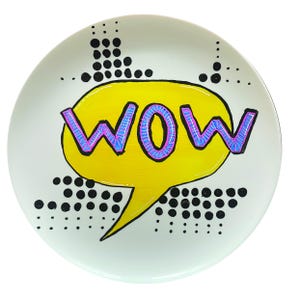 May include: A white ceramic plate featuring a yellow speech bubble with the word "WOW" in purple and blue outlined letters. Black dots and dashes surround the bubble, in a comic book style.