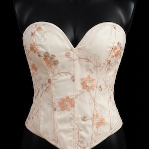 May include: Ivory-colored corset with floral embroidery. The strapless bodice features a sweetheart neckline and is adorned with delicate peach and gold-toned embroidered flowers and vines. The corset has a structured design.
