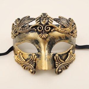 Puede incluir: Una máscara veneciana dorada y negra con un acabado envejecido y desgastado. La máscara presenta elementos decorativos que incluyen una cara central, hojas de laurel y figuras aladas. Se adjunta una cinta negra para llevarla.