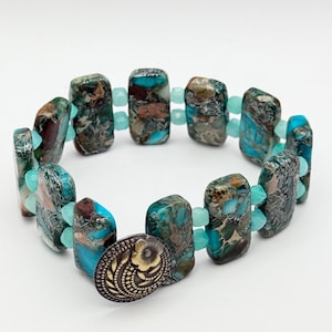 May include: A bracelet featuring rectangular beads with a marbled pattern in shades of teal, brown, and black, accented by small, faceted light blue beads. The piece is secured with a round, metallic button clasp featuring an embossed floral design.