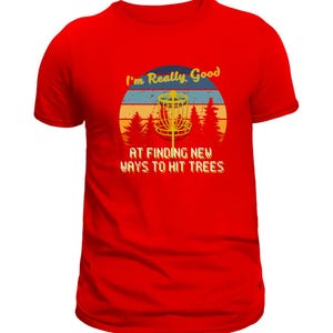 Op de afbeelding: Rood t-shirt met een grafisch ontwerp voor discgolf liefhebbers. Het ontwerp toont een discgolfmand, bomen en de tekst "I'm Really Good At Finding New Ways To Hit Trees" in retro stijl.