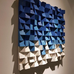 Puede incluir: Una moderna obra de arte mural compuesta por bloques de madera en diferentes tonos de azul y blanco. Los bloques están dispuestos para crear un efecto texturizado y tridimensional, con un degradado de azul oscuro en la parte superior a blanco en la inferior. La obra está montada en una pared.